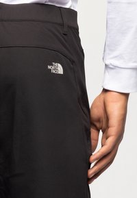 Gros plan d'une personne portant un pantalon noir The North Face avec une chemise blanche à manches longues, main reposant près de la poche latérale.