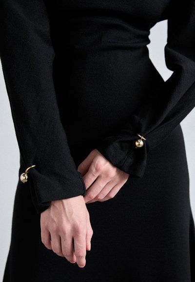 Black long-sleeve dress with fitted silhouette. Sleeves feature a knotted detail at the wrist with a gold spherical accent. Soft texture.