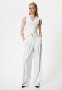Weiße gestreifte ärmellose Bluse mit goldenen Knöpfen und passenden weit geschnittenen Hosen, die eine hohe Taille und ein plissiertes Design aufweisen.