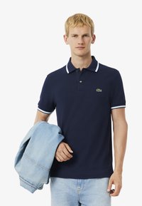 Camisa polo azul marino con ribetes blancos en el cuello y las mangas, que presenta un logotipo bordado en verde. Chaqueta de mezclilla clara colgada del brazo.