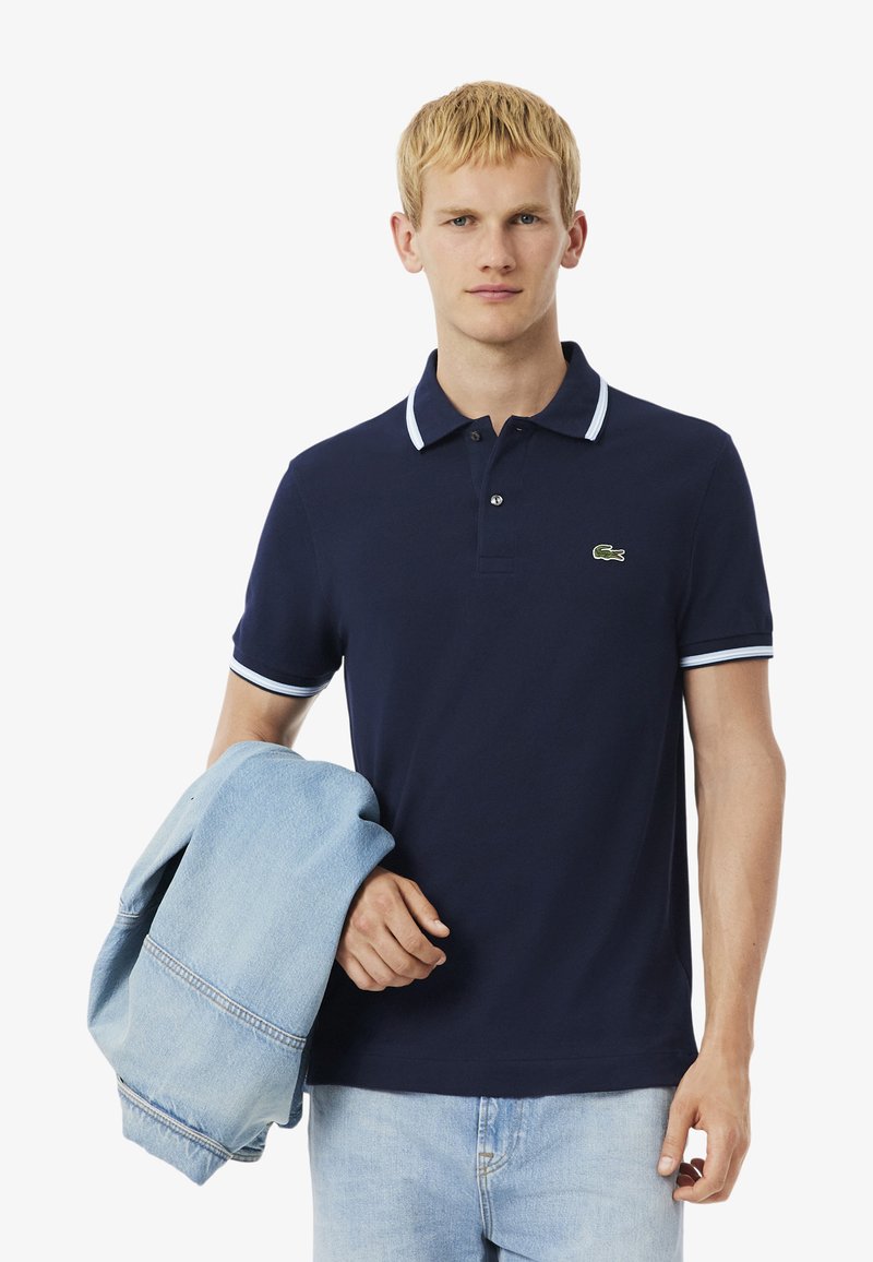 Camisa polo azul marino con ribetes blancos en el cuello y las mangas, que presenta un logotipo bordado en verde. Chaqueta de mezclilla clara colgada del brazo.