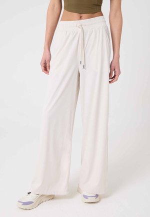 Persona con pantalones blancos sueltos de tiro alto con cordón y zapatillas, de pie contra un fondo claro y liso.