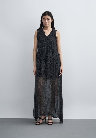 Γυναίκα στέκεται φορώντας αμάνικο μακρύ μαύρο διάφανο φόρεμα με μικρές λευκές βούλες και μαύρα πέδιλα με τακούνια σε απλό φόντο.