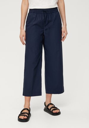 Vrouw draagt marineblauwe wijde cropped broek met elastische tailleband en zwarte sandalen, staand op een lichte achtergrond.