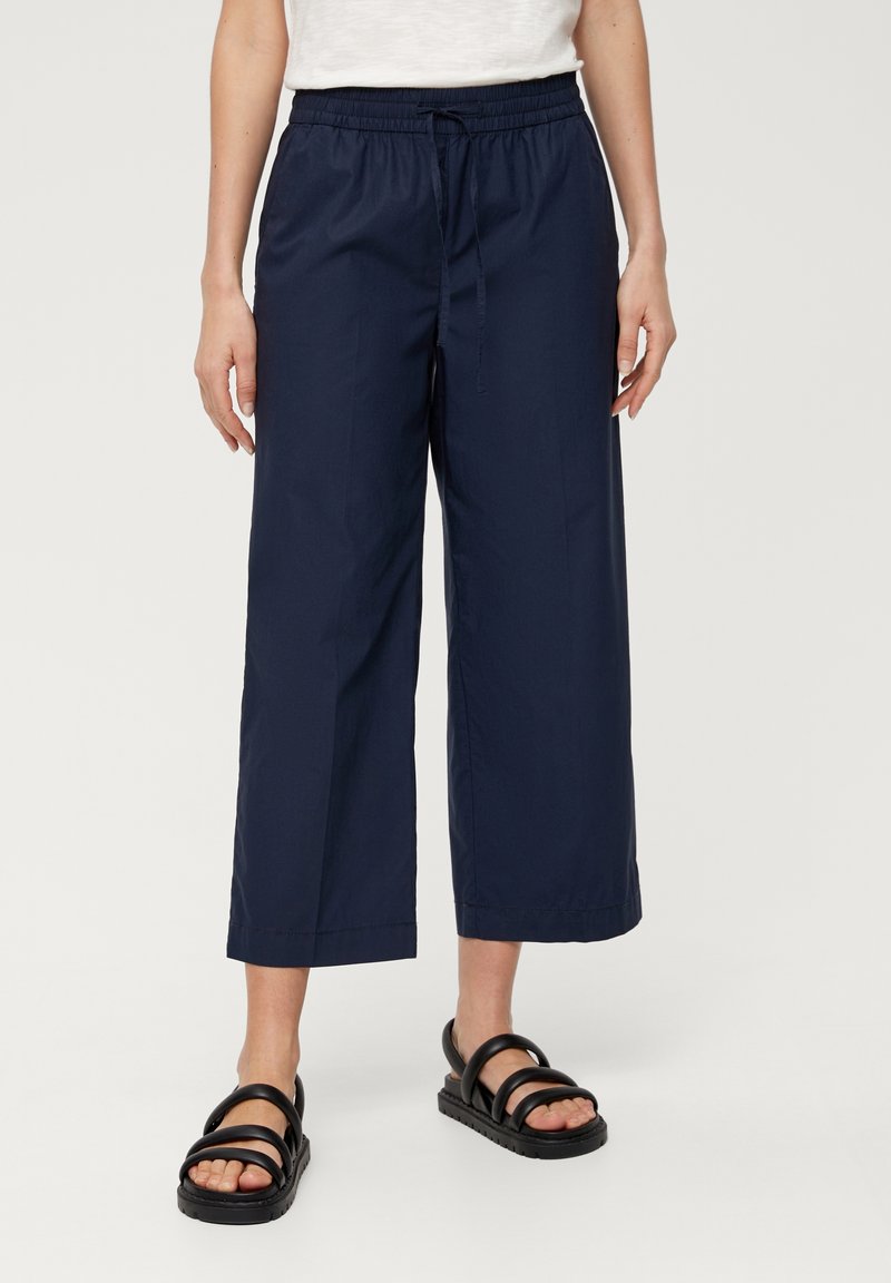 Femme portant un pantalon court large bleu marine avec une ceinture élastique et des sandales noires, debout sur un fond clair.