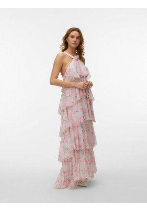 Femme portant une robe maxi rose à fleurs sans manches et superposée, à col licou, debout les mains croisées et regardant devant elle.