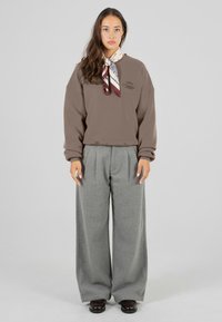 Brun genser med logo, kombinert med vide grå bukser og et mønstrete skjerf. Glatt stoff og avslappet passform med store ermer.