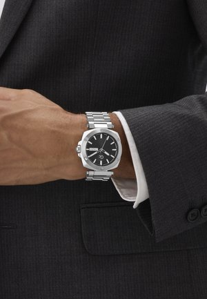 Uomo in completo scuro che sistema un orologio da polso argentato con quadrante nero e cinturino in metallo, con indicazione del giorno e della data sul quadrante.