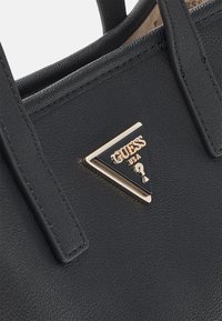 Sort syntetisk håndtaske med struktureret overflade, dobbelte håndtag og et trekantet Guess-logo med gulddetaljer på fronten.