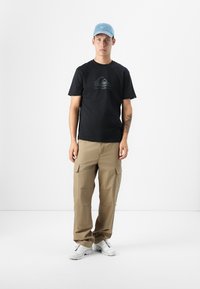 Ungdommelig mannlig modell står i en minimalistisk setting, iført en svart t-skjorte, khaki cargobukser og en lyseblå caps.