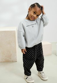 Svetlosivý sweatshirt s nápisom "HAPPY LITTLE MIND", čierne kárované nohavice a béžové tenisky. Má voľný strih a jemnú textúru.