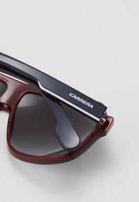 Ochelari de soare roșii și navy cu un finisaj lucios, lentile gri colorate, design de cadru îndrăzneț și logoul "CARRERA" pe tâmplă.