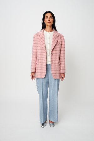 Γυναίκα με ροζ καρό σακάκι, λευκό πουκάμισο με κουμπιά, μπλε παντελόνι φαρδύ με ρίγες και ασημένια παπούτσια, στέκεται μπροστά σε λευκό φόντο.