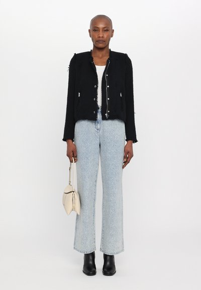 Schwarze Texturjacke mit fransigen Kanten, silbernen Knöpfen; kombiniert mit hellblauen geraden Jeans und schwarzen Stiefeletten. Weiße Handtasche.