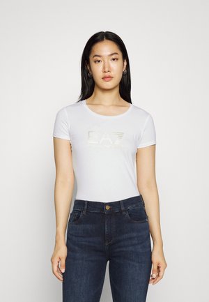 Maglietta bianca in cotone con scollatura rotonda, caratterizzata da una grafica con logo dorato sulla parte anteriore. Abbinata a jeans blu scuro, di trama liscia.