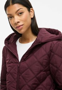 Burgundy quilted jacket with a zip front and hood, featuring a diamond pattern and smooth texture, worn over a white top.