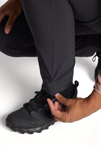 Schwarze leichte Hosen mit verstellbaren Bündchen werden über schwarzen Sportschuhen befestigt. Der Stoff wirkt glatt und langlebig.