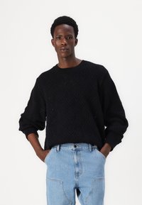 Sort strikket sweater med et diamantmønster, ribbet hals og manchetter. Kombineret med lyseblå denim jeans. Modellen står med hænderne i lommerne.
