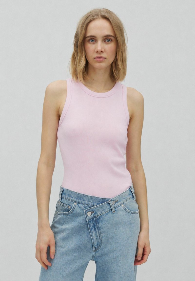 Femme aux cheveux blonds jusqu'aux épaules, vêtue d'un haut rose clair sans manches et d'un jean bleu ample, debout devant un fond uni clair.