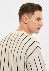 Kremowy sweter z dzianiny z czarnymi pionowymi paskami, ściągaczowym kołnierzem i metką z tyłu. Luźny krój i fakturowany materiał.