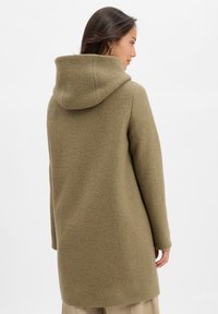 Frau mit langen dunklen Haaren, die einen olivgrünen, kapuzenbehangenen Wollmantel trägt und leicht zur Seite gedreht vor einem weißen Hintergrund steht.