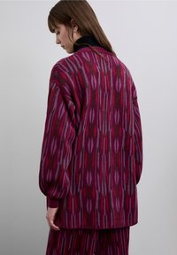 Knitted cardigan in shades of purple and red with a geometric pattern. Features a relaxed fit, bold sleeves, and a high collar. Textured finish.