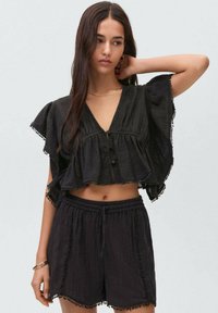 Čierny cropped top s volánovými rukávmi a výstrihom do V, s gombíkovým zapínaním. Skombinovaný s čiernymi šortkami s ozdobným okrajom.