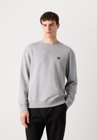Sudadera gris claro de textura suave con mangas largas y cuello redondo, que presenta un pequeño parche negro con el logo en la parte superior izquierda.
