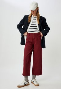 Wijd-lange bordeauxkleurige broek, gestreept wit en marineblauw shirt, zwarte blazer en een beige pet. Witte sneakers met zwarte accenten en grijze sokken.