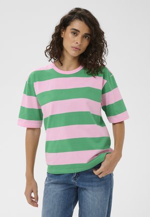 Młoda osoba o kręconych ciemnych włosach, ubrana w luźną poziomo paskowaną różowo-zieloną koszulkę i niebieskie dżinsy, na neutralnym tle.