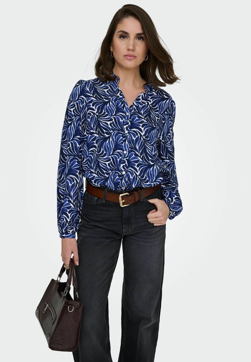 Femme portant un chemisier à motifs de feuilles bleu et blanc, un jean noir avec ceinture marron, tenant un sac à main marron foncé, debout devant un fond uni.