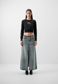 Sudadera corta negra con un corte, combinada con una falda de mezclilla larga y desgastada que presenta una abertura en la parte frontal y una cinturilla con acentos rojos. Botas negras.