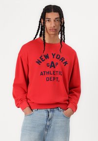 Sudadera roja con texto en marino que dice "NUEVA YORK GAP DEPARTAMENTO DE ATLETISMO". Puños y dobladillo acanalados, corte relajado, usada con jeans de color azul claro.