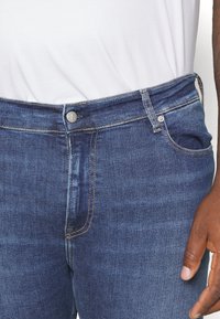 Tmavě modré džíny s klasickým střihem, s jemným stříbrným knoflíkem, zipovým zapínáním a kontrastním zlatým prošíváním. Viditelná boční kapsa.