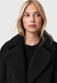 Černý texturovaný kabát s klopovým límcem, vyrobený z nadýchaného materiálu. Nosený přes černý svetr, zdůrazňující teplé a útulné designové prvky.