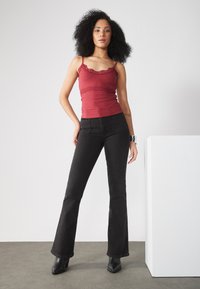 Rød ribbeformet topp med blonder, kombinert med svarte flared jeans og svarte ankelboots med hæler. Modellen står foran en nøytral bakgrunn.
