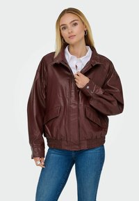 Giacca in pelle bordeaux con un taglio rilassato, dotata di due tasche frontali, polsini con bottone e orlo elastico. Indossata sopra una camicia bianca.