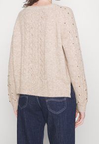 Beige gebreide trui met een gestructureerd patroon, comfortabele pasvorm en opgerolde randen. Heeft decoratieve gouden accenten op de mouwen.