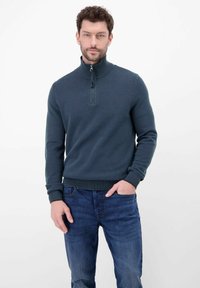 Granatowy sweter na zamek wykonany z teksturowanej dzianiny, z wysokim kołnierzem, ściągaczami na mankietach i w pasie. Połączony z niebieskimi dżinsami.