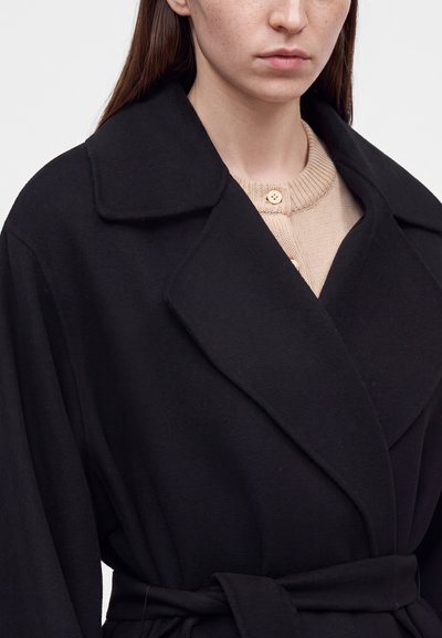 Femme portant un manteau noir ceinturé par-dessus un pull beige boutonné, avec col et haut du torse visibles sur un fond uni.
