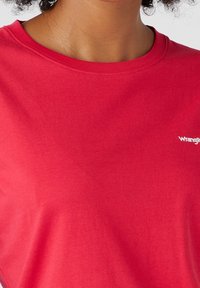Červené bavlněné tričko s kulatým výstřihem, na hrudi s bílým logem "Wrangler", hladká textura a klasický střih.