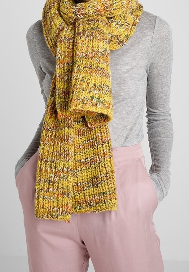 Écharpe tricotée jaune avec une texture multicolore. Conception épaisse et volumineuse avec un motif côtelé, drapée autour du cou. Portée avec un haut gris et un pantalon rose.