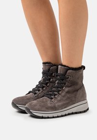 Šedé semišové kotníkové boty s černými tkaničkami a umělou kožešinou, které mají robustní bílou podrážku a kovové očko. Texturovaný materiál dodává hloubku.