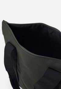 Mørkegrønn totebag med en glatt ytre og svarte tekstilremmer. Den glidelås åpningen avdekker et svart interiør med en lomme.