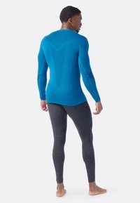 Man standing barefoot facing backward wearing a blue long-sleeve top and dark gray fitted leggings against a plain background.