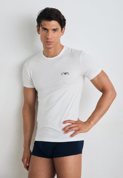 Hvid bomulds T-shirt med korte ærmer, rund hals og lille marineblå logo på venstre bryst. Matchet med mørkeblå shorts. Glat tekstur.