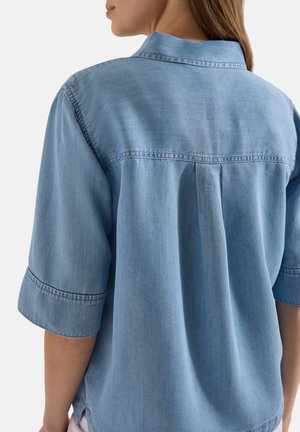 Kvinde iført løs, lyseblå denimskjorte med albuelange ærmer, set bagfra med synlige detaljer ved skulderstykke og central læg.