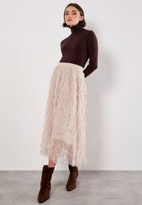 Un pull à col roulé marron à manches longues associé à une jupe plissée légère de couleur rose pâle, arrivant au genou. Le mannequin porte des bottines marron. Textures douces et lisses en évidence.