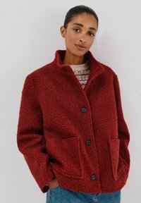 Rote, strukturierte Fleecejacke mit leicht oversized Passform, schwarzen Knöpfen, zwei Vordertaschen und einem Kragen. Getragen über einem gemusterten Pullover.