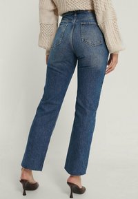 Mujer lleva puesta unos vaqueros de tiro alto de color azul con bolsillos traseros, dobladillo sin rematar, suéter de punto beige y zuecos marrones de tacón alto.